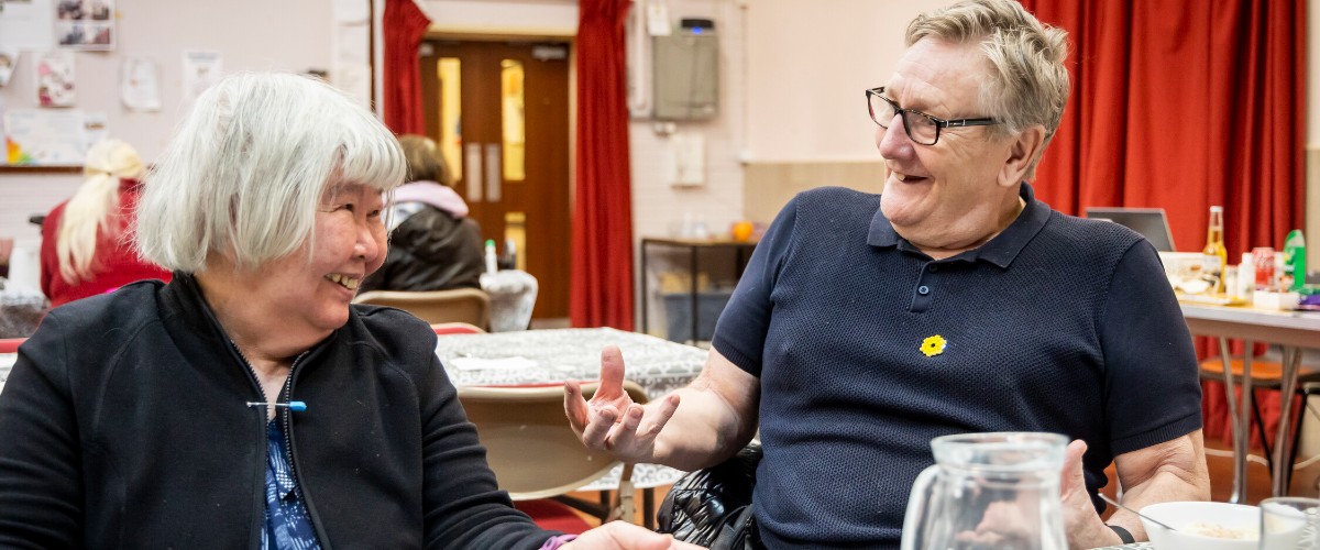 a man and a woman sat together, chatting, in a community hall